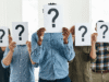 Business Valuation in 2026: How to Know What Your Company Is Worth Group of people in a bright office holding white signs with large question marks, obscuring their faces for anonymity or uncertainty.