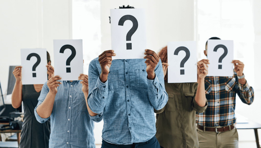 Group of people in a bright office holding white signs with large question marks, obscuring their faces for anonymity or uncertainty.