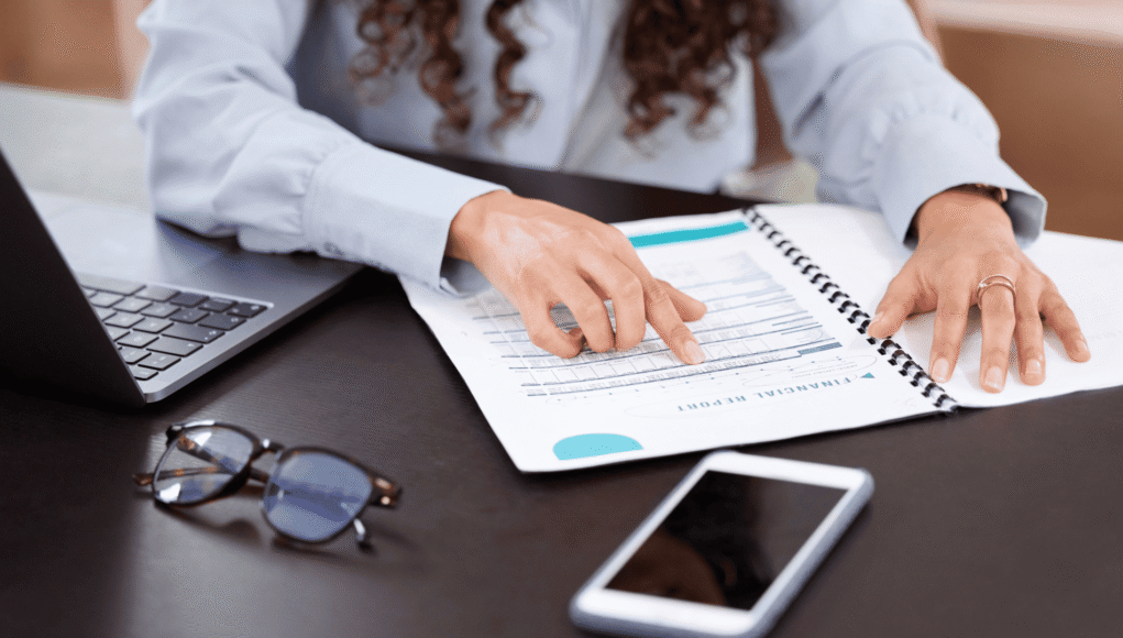Person points to a financial report in a spiral notebook on a desk, with a laptop nearby.