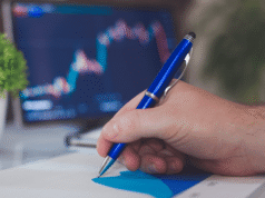 Dental Practice Overhead in 2026: Benchmarks & Profit Optimization Guide Close-up of a hand writing with a blue ballpoint pen on white paper, with a blurred laptop in the background