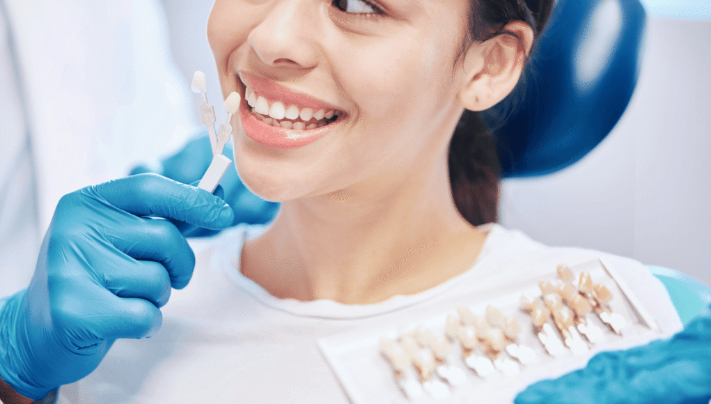 Dentist in blue gloves holds tooth shade guide next to patient's smile to match tooth color.