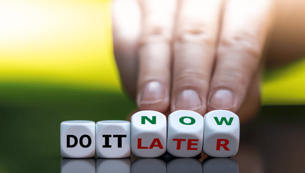 Hand placing blocks that spell 'DO IT LATER' with 'NOW' visible, conveying procrastination.