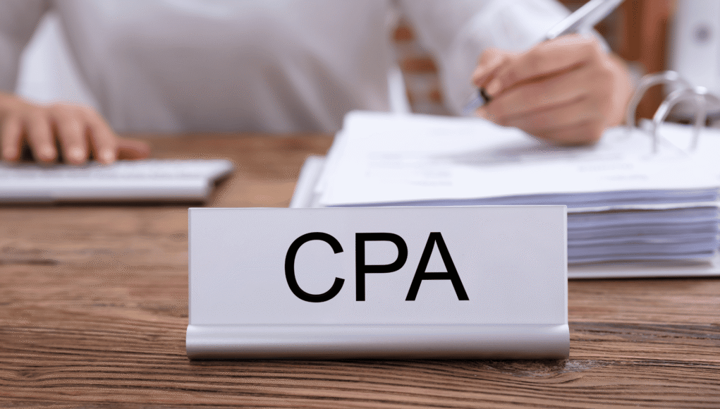 Close-up of a CPA nameplate on a desk with a stack of papers and a person writing in the background.