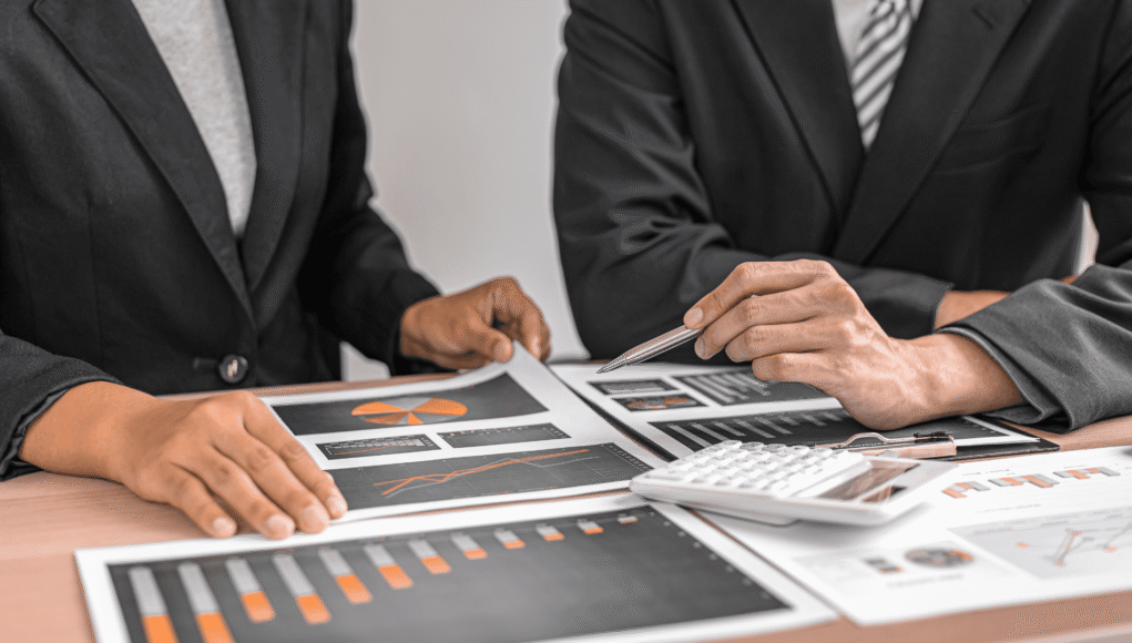 Two professionals in suits review financial charts and graphs spread across a desk, using a pen and calculator nearby.