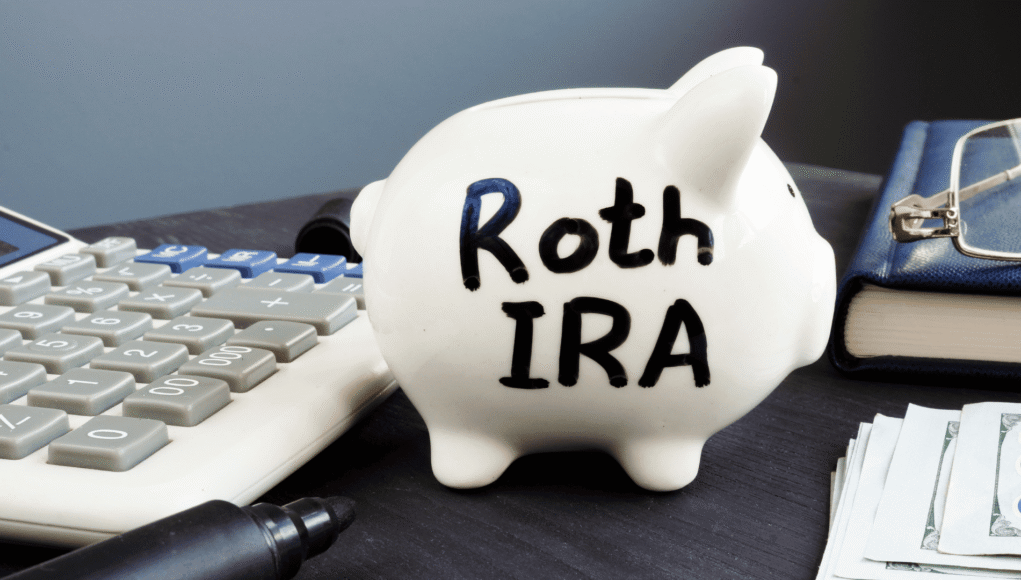 White piggy bank labeled Roth IRA on a desk next to a calculator, pen, wallet, and stacks of cash—conveys retirement savings.