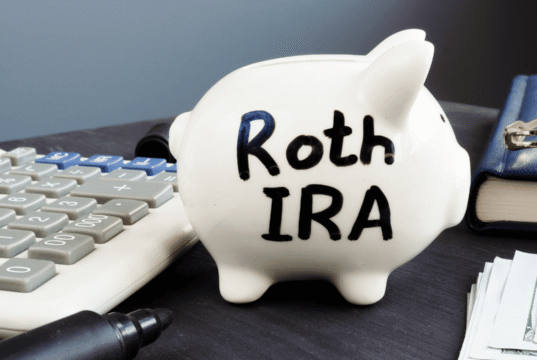 Roth Conversion Strategy 2026: How High-Income Earners Can Save on Taxes White piggy bank labeled Roth IRA on a desk next to a calculator, pen, wallet, and stacks of cash—conveys retirement savings.