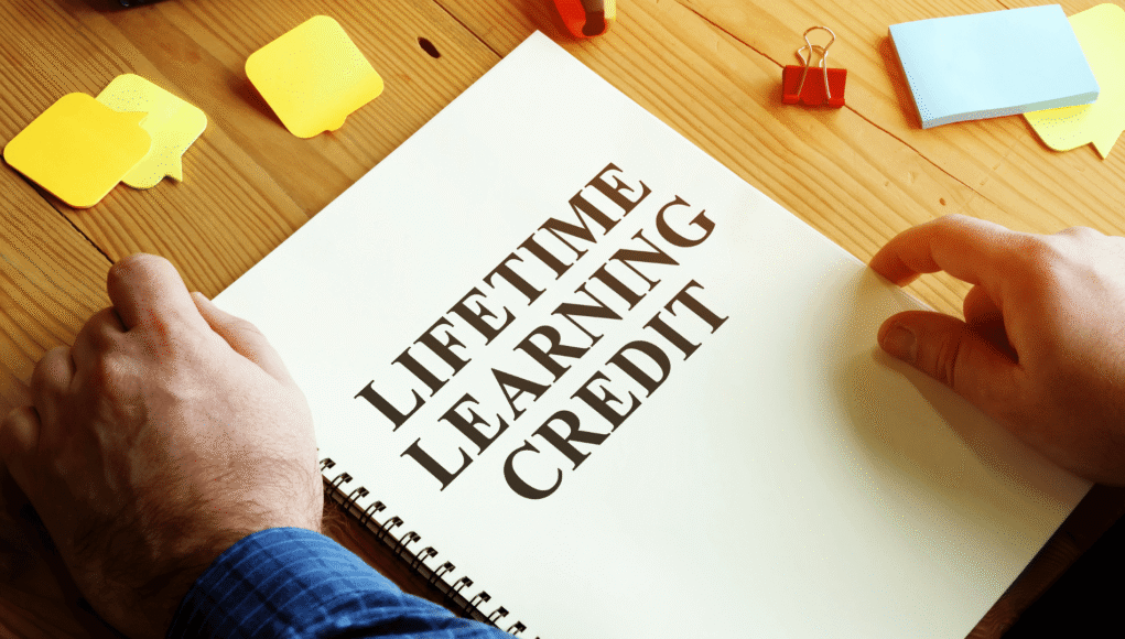 Notebook open to a page titled 'Lifelong Learning Credit' with hands resting on the page and yellow sticky notes on a wooden desk.