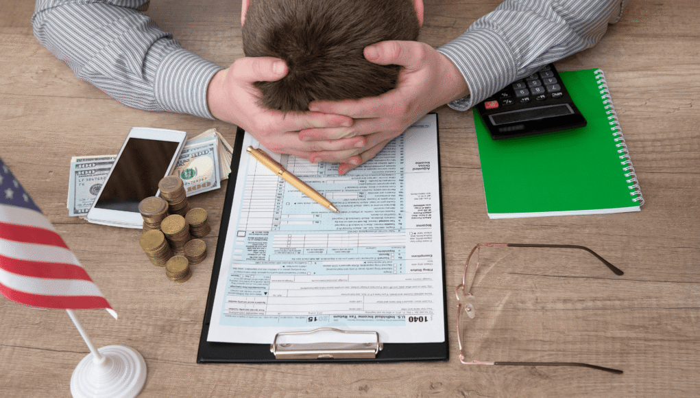 Person rests head in hands over a tax form on a clipboard, surrounded by money, phone, and calculator in a stressful office scene commonly linked to finances.